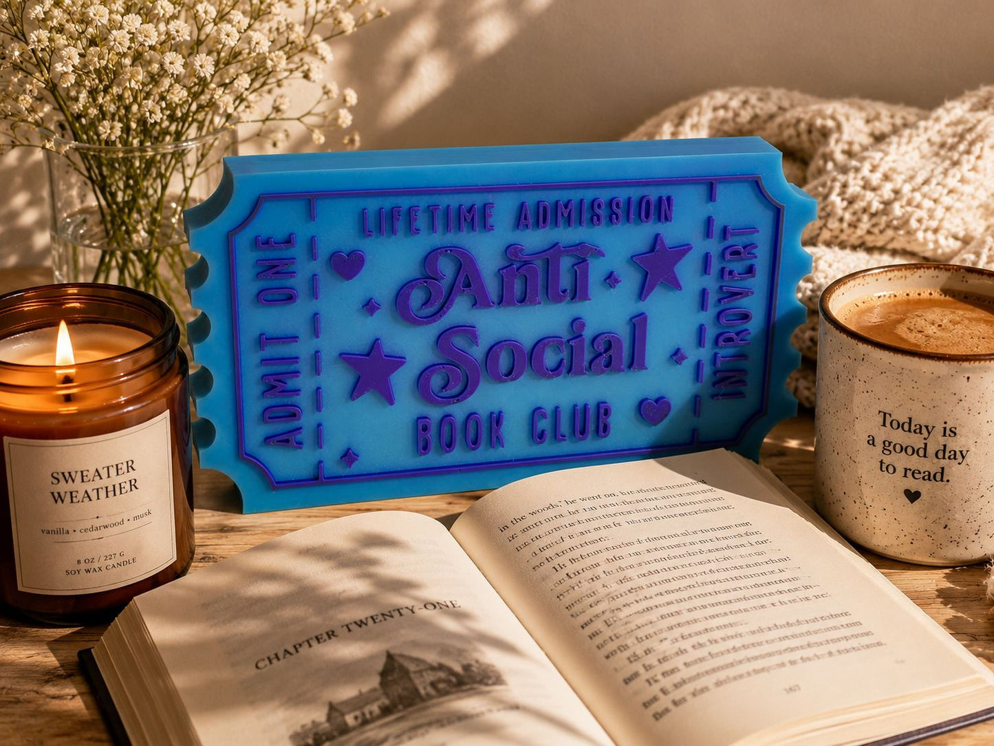a cozy scene with a blue ticket-shaped sign for the Anti-Social Book Club, a candle, a mug of coffee, and an open book on a wooden surface.