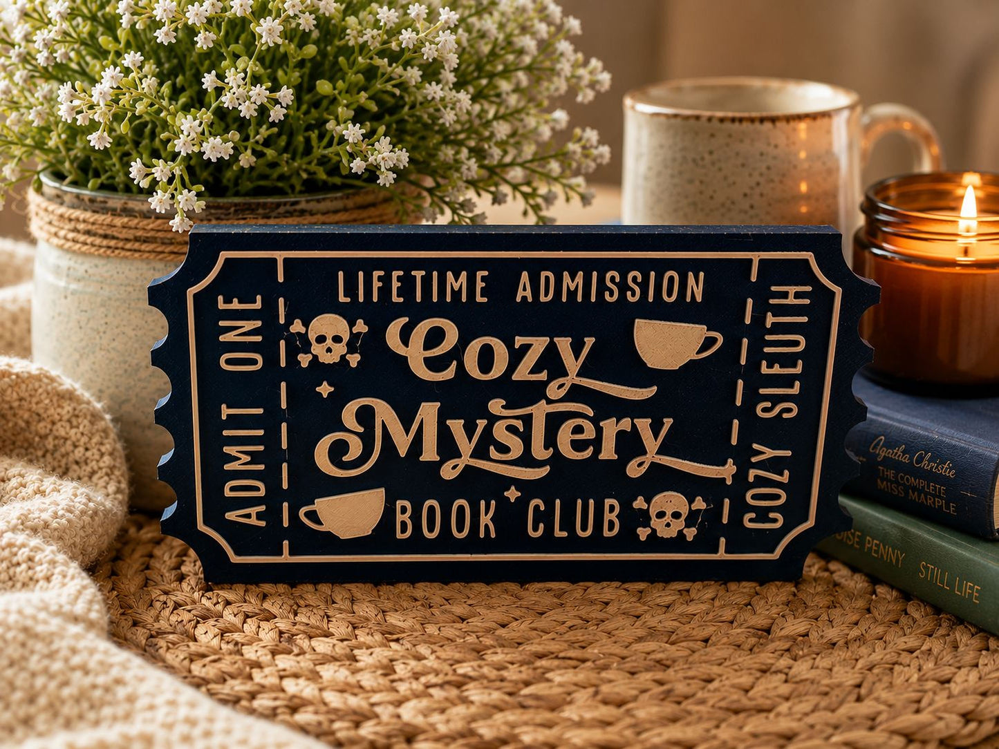 a blue ticket-shaped sign with the words "Lifetime Admission Cozy Mystery Book Club" written on it, placed on a woven surface. There are also other items in the scene, such as a candle, a mug, and a book, creating a cozy and inviting atmosphere.