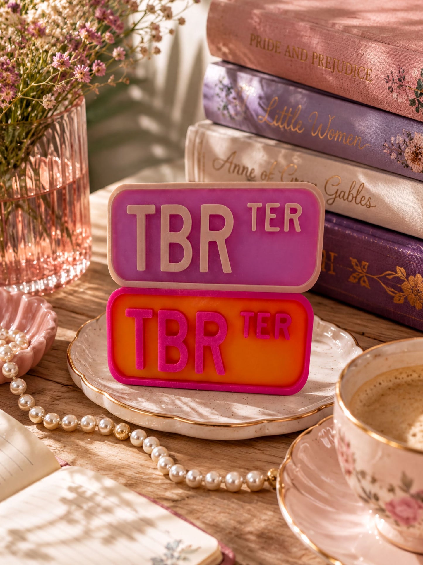 a pink and orange "TBR" sign, a stack of books, a teacup, pearls, and a vase with flowers on a wooden surface.