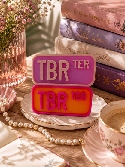 a pink and orange "TBR" sign, a stack of books, a teacup, pearls, and a vase with flowers on a wooden surface.