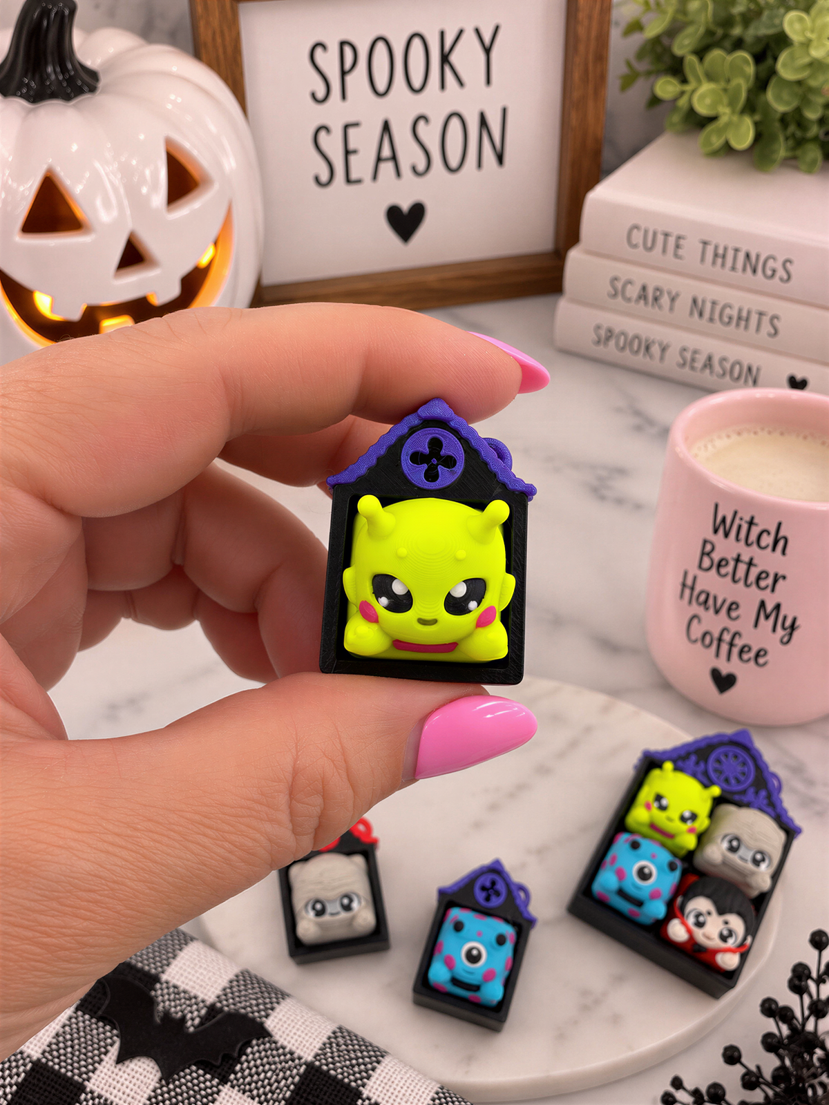A hand holding a small, colorful keychain with a cartoon character on it, surrounded by Halloween decorations and a mug with a witch-themed design.