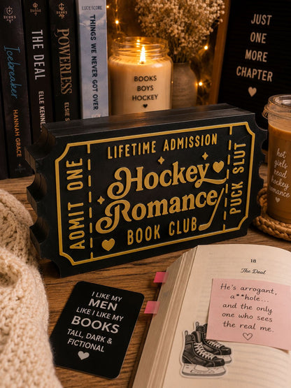 a collection of items related to hockey romance, including a ticket-shaped object, a book, and a note. The items are arranged on a wooden surface, with a candle and a mug visible in the background.