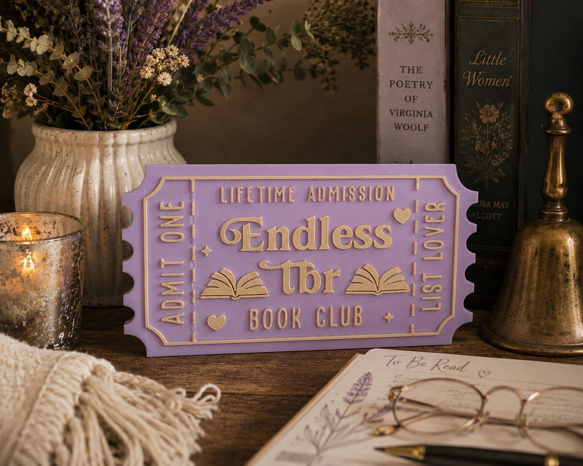 a purple ticket-shaped sign for a book club, surrounded by various books, a vase of flowers, and a candle on a wooden surface.