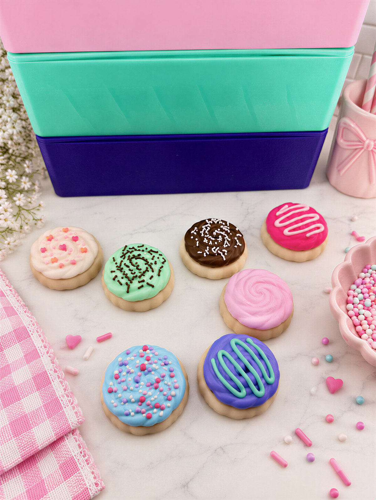 a variety of colorful and decorated cookies, including sugar cookies with pink frosting, chocolate cookies with sprinkles, and cookies with blue and green frosting. The cookies are arranged on a marble surface, with some scattered around and a pink container visible in the background.