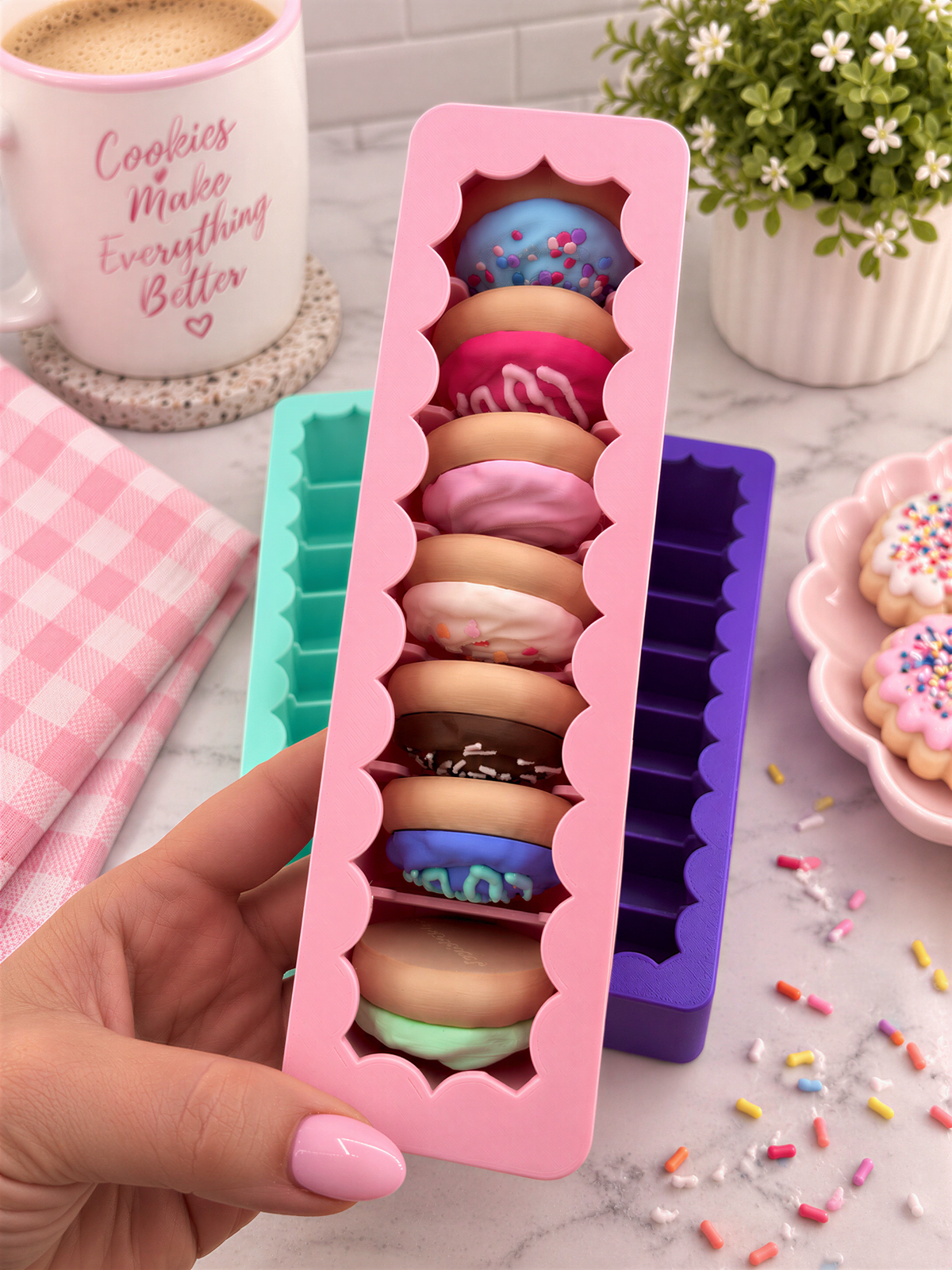 A hand holding a pink container filled with colorful donut-shaped cookies, surrounded by other donut-shaped cookies and a mug with the text "Cookies Make Everything Better".